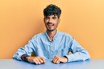 Young hispanic man wearing casual clothes sitting on the table winking looking at the camera with sexy expression, cheerful and happy face.