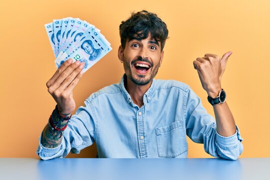 Young hispanic man holding yuan chinese banknotes sitting on the table pointing thumb up to the side smiling happy with open mouth