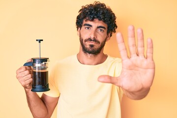 Handsome young man with curly hair and bear holding french coffee maker with open hand doing stop sign with serious and confident expression, defense gesture