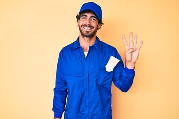 Handsome young man with curly hair and bear wearing builder jumpsuit uniform showing and pointing up with fingers number four while smiling confident and happy.