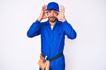 Handsome young man with curly hair and bear weaing handyman uniform trying to open eyes with fingers, sleepy and tired for morning fatigue