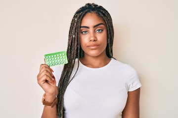 Young african american woman with braids holding birth control pills thinking attitude and sober...