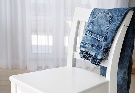 Blue denim jeans stacked on white chair with tulle window on background with free place for your product advertising. Casual clothes or laundry concept with copy space.