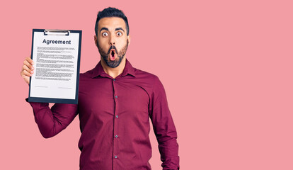 Young hispanic man holding clipboard with agreement document scared and amazed with open mouth for surprise, disbelief face