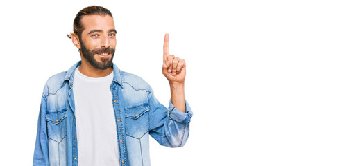 Attractive man with long hair and beard wearing casual denim jacket showing and pointing up with finger number one while smiling confident and happy.