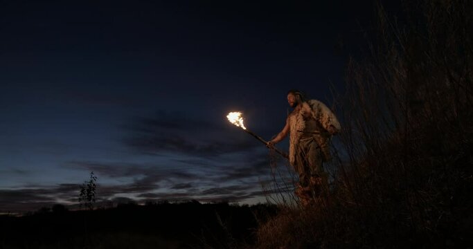 Primitive hunter gatherer wearing sheepskins and carrying a bow and arrows walking by torch light using a burning stick taper at night in countryside. Portrait of primeval caveman wearing animal night