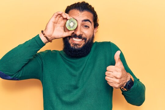 Young arab man holding kiwi smiling happy and positive, thumb up doing excellent and approval sign