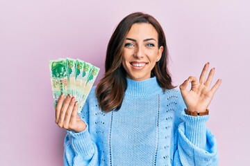 Young brunette woman holding hong kong 50 dollars banknotes doing ok sign with fingers, smiling friendly gesturing excellent symbol