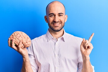 Young handsome bald man holding brain smiling happy pointing with hand and finger to the side