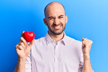 Young handsome bald man holding heart screaming proud, celebrating victory and success very excited with raised arm