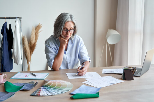 Senior mature elegant female fashion designer drawing sketches in studio office. Middle aged stylish grey-haired older woman small business owner creating new fashion design clothes in atelier.