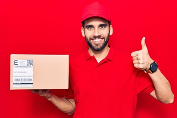 Young handsome man with beard wearing delivery uniform holding package smiling happy and positive, thumb up doing excellent and approval sign