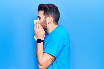 Young hispanic man illness using paper handkerchief on nose. Standing over isoltated blue background