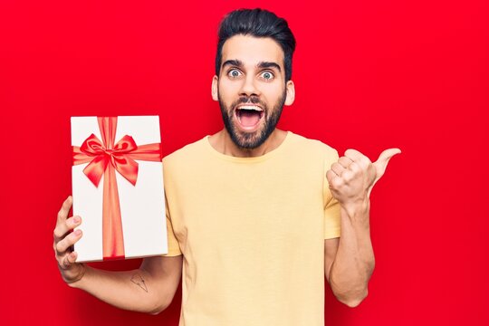 Young handsome man with beard holding gift pointing thumb up to the side smiling happy with open mouth