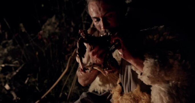 Ravenous primitive man clad in sheepskins squatting alongside a burning fire at night tearing barbecued meat off the bone with his teeth in close up. Close up portrait caveman eating in at night.