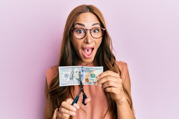 Young brunette woman cutting dollars with scissors for currency devaluation celebrating crazy and amazed for success with open eyes screaming excited.