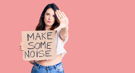 Beautiful young brunette woman holding make some noise banner with open hand doing stop sign with serious and confident expression, defense gesture