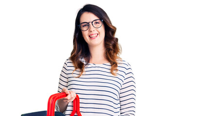 Beautiful young brunette woman holding supermarket shopping basket looking positive and happy standing and smiling with a confident smile showing teeth