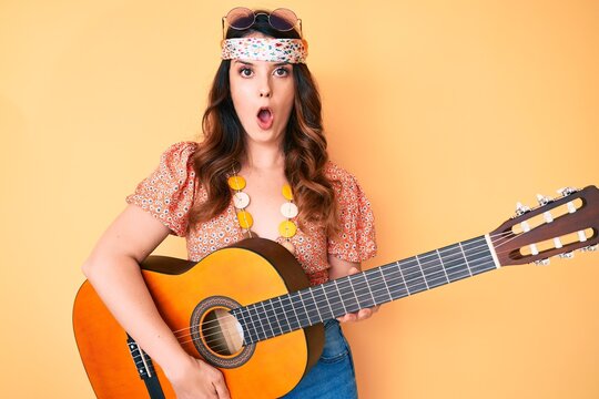 Young beautiful brunette woman wearing boho style playing classical guitar scared and amazed with open mouth for surprise, disbelief face
