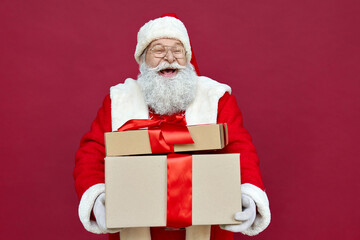 Happy funny old bearded Santa Claus wears costume holds many presents, Merry Christmas giftboxes wrapped with red ribbon, laughing standing with gift boxes having fun isolated on red xmas background.