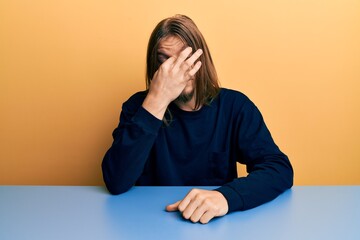 Handsome caucasian man with long hair wearing casual clothes sitting on the table tired rubbing...