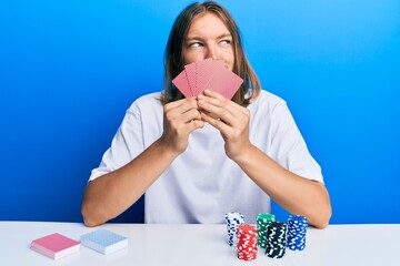 Handsome caucasian man with long hair playing gambling poker covering face with cards smiling looking to the side and staring away thinking.