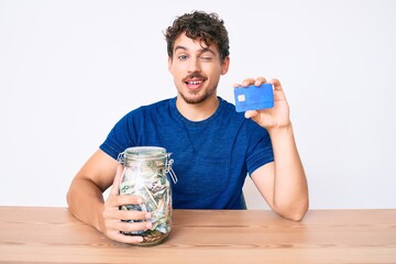 Young caucasian man with curly hair holding credit car and jar with dollars winking looking at the...