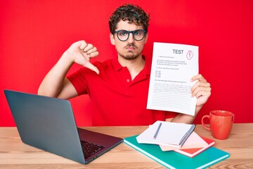 Young caucasian man with curly hair sitting on the table showing failed exam with angry face, negative sign showing dislike with thumbs down, rejection concept