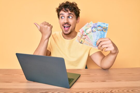 Young caucasian man with curly hair sitting on the table working with laptop and holding canadian dollars banknotes pointing thumb up to the side smiling happy with open mouth