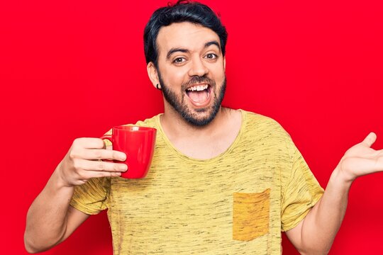 Young hispanic man holding coffee celebrating achievement with happy smile and winner expression with raised hand