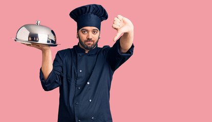 Young hispanic man wearing cooker uniform holding tray with angry face, negative sign showing...