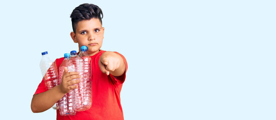 Little boy kid holding recycling plastic bottles pointing with finger to the camera and to you, confident gesture looking serious