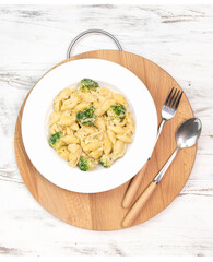 Pasta with green vegetables, broccoli, melted cheese and creamy sauce in white bowl on white table.