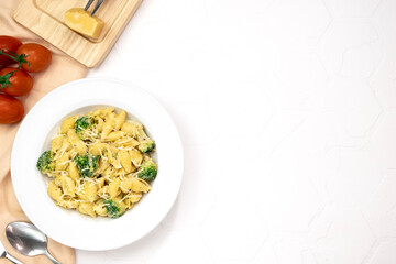 Pasta with green vegetables, broccoli, melted cheese and creamy sauce in white bowl on white table.
