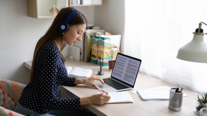 Side view focused young beautiful caucasian woman wearing wireless headphones, studying remotely using laptop at home office, smart millennial female student involved in research article writing.