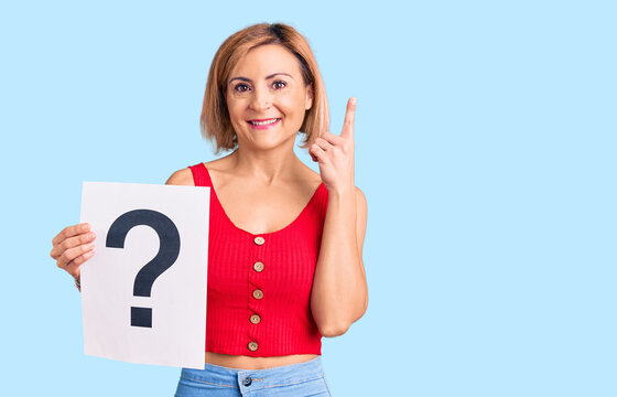 Young blonde woman holding question mark surprised with an idea or question pointing finger with happy face, number one