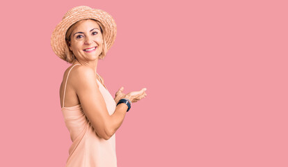 Young blonde woman wearing summer hat pointing aside with hands open palms showing copy space, presenting advertisement smiling excited happy