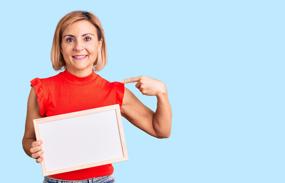 Young blonde woman holding empty white chalkboard pointing finger to one self smiling happy and proud