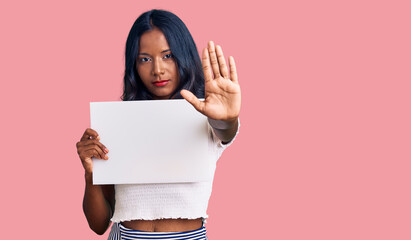 Young indian girl holding blank empty banner with open hand doing stop sign with serious and...