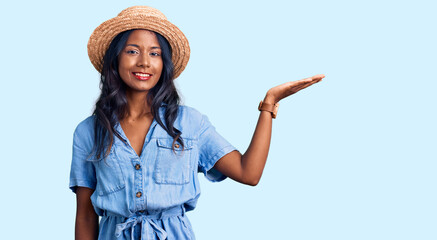Young indian girl wearing summer hat smiling cheerful presenting and pointing with palm of hand...