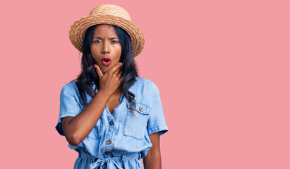 Young indian girl wearing summer hat looking fascinated with disbelief, surprise and amazed expression with hands on chin