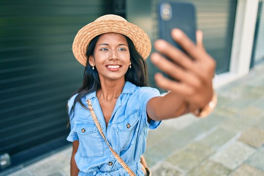 Young beautiful indian woman wearing summer hat smiling happy making selfie by the smartphone at the city.