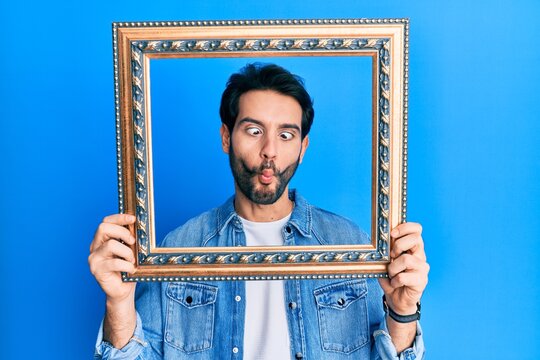 Young hispanic man holding empty frame making fish face with mouth and squinting eyes, crazy and comical.