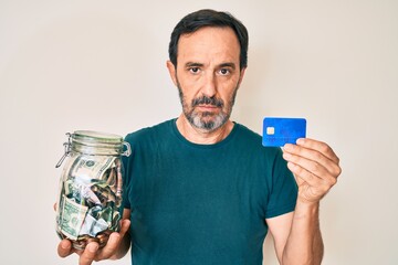 Middle age hispanic man holding credit car and jar with dollars relaxed with serious expression on face. simple and natural looking at the camera.