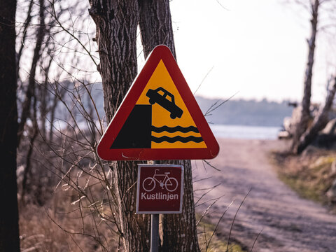 Warning Sign Depicting A Car Falling Over An Edge Into Water Next To A Sign Marking The Way For The Cycling Trail Kustlinjen