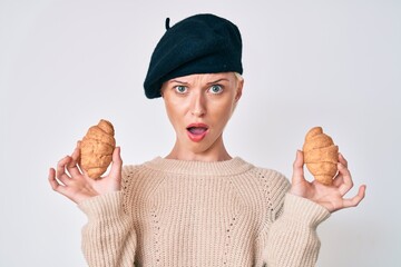 Young caucasian woman wearing french look with beret holding croissants in shock face, looking skeptical and sarcastic, surprised with open mouth