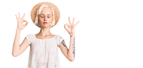 Young blonde woman with tattoo wearing summer hat relax and smiling with eyes closed doing meditation gesture with fingers. yoga concept.