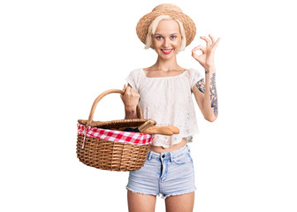 Young blonde woman with tattoo wearing summer hat and holding picnic wicker basket with bread doing ok sign with fingers, smiling friendly gesturing excellent symbol