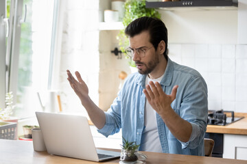 Frustrated angry young man in eyeglasses looking at laptop screen, feeling nervous about bad device work or poor internet connection in kitchen. Unhappy shocked guy reading message with bad news.