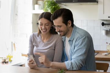 Obraz premium Pretty young woman holding smartphone in hands, showing funny photo editing application or online game to smiling handsome man, using electronic device together at free leisure time at home.